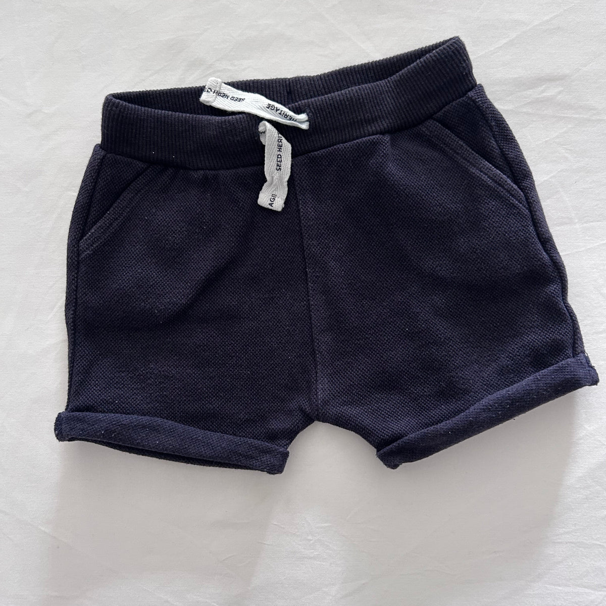 A pair of dark navy blue knit shorts with ribbed cuffs, laid flat on a white textured fabric background. A small white paper tag is attached near the waistband.