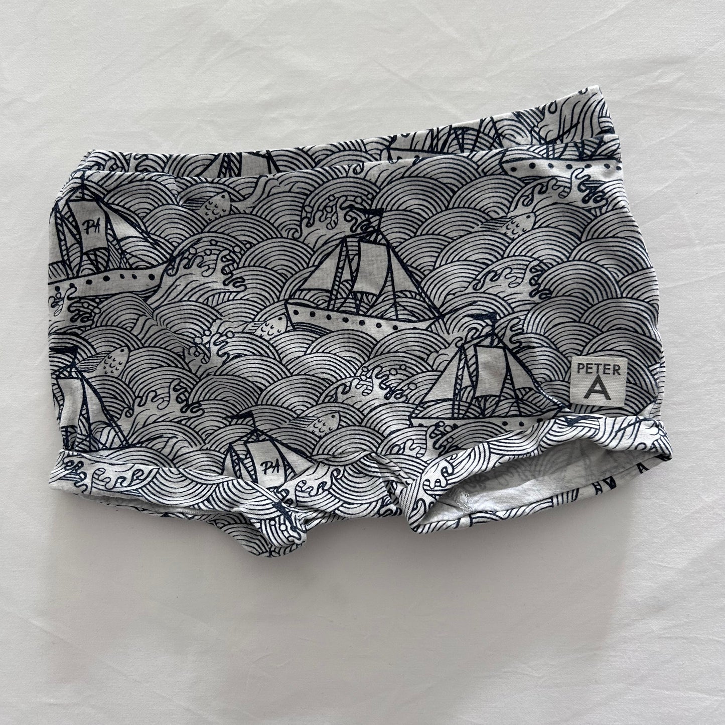 A pair of shorts with a navy blue and white wave and sailboat pattern, laid flat on a white fabric background.