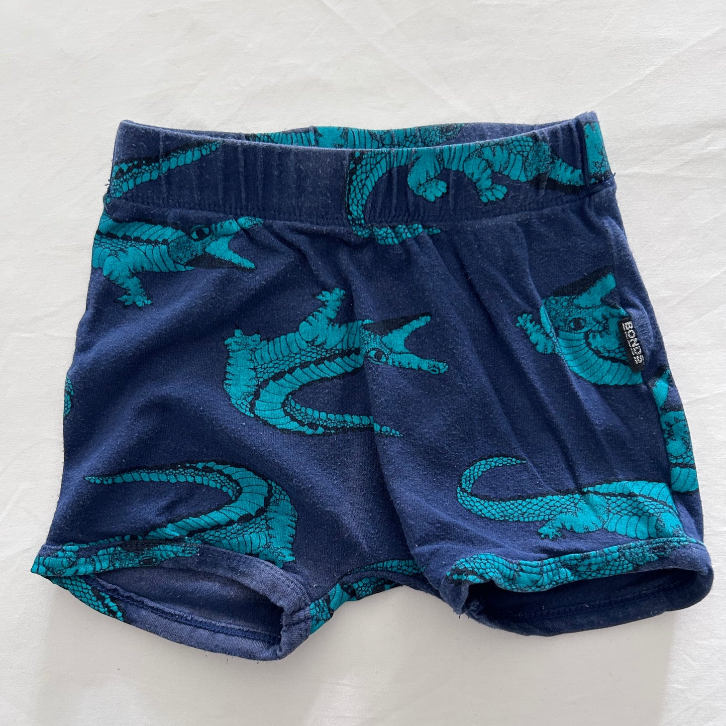 A pair of toddler boy's shorts in navy blue with a teal alligator print, laid flat on a white fabric background.