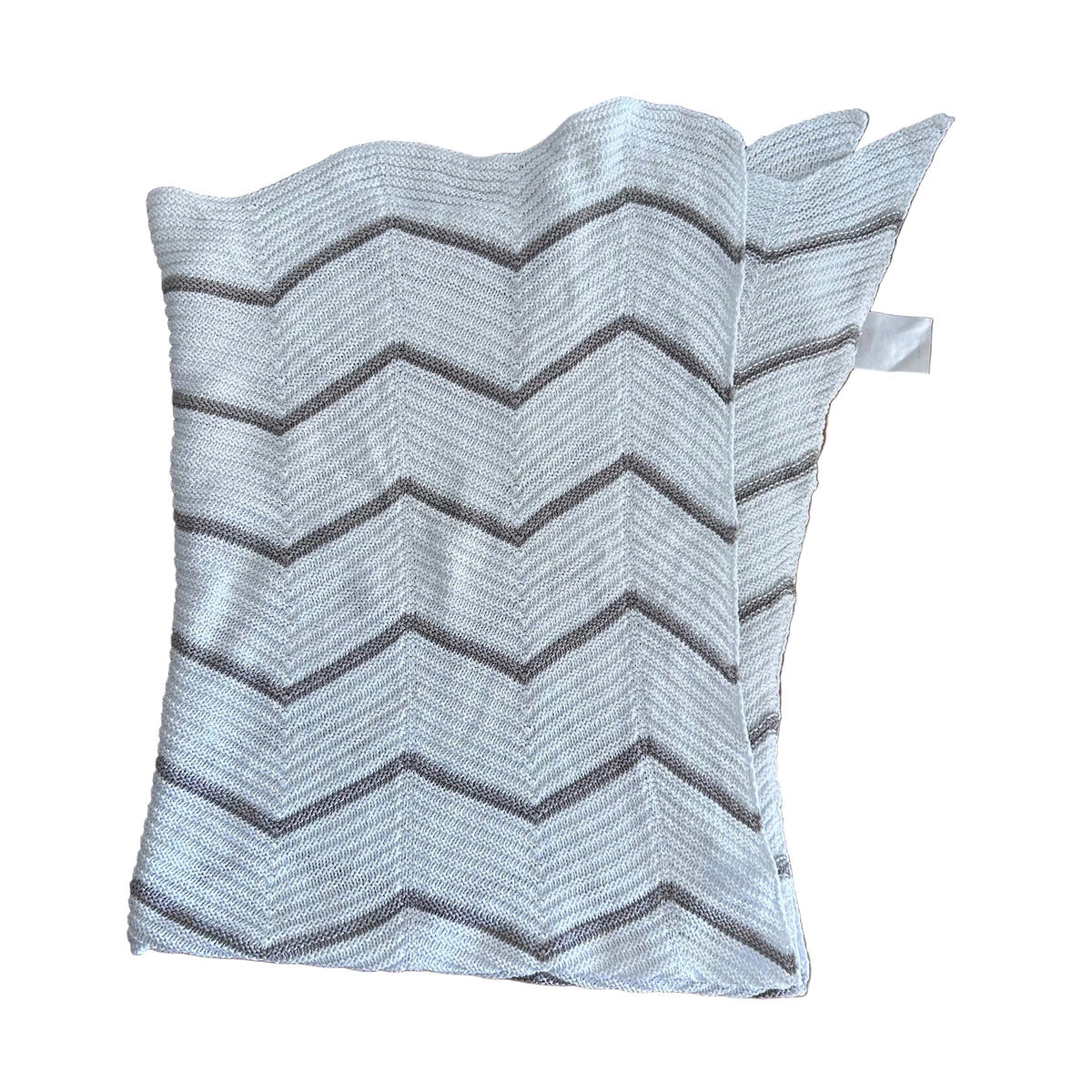 A white knit baby blanket with a gray chevron pattern, laid flat on a wooden table. A small white fabric tag is visible on the right side, and a person's fingers are visible in the bottom right corner.