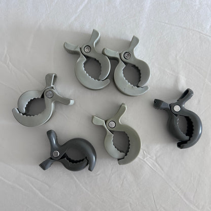 Six plastic spring clamps in various shades of gray and charcoal, arranged on a white fabric background.