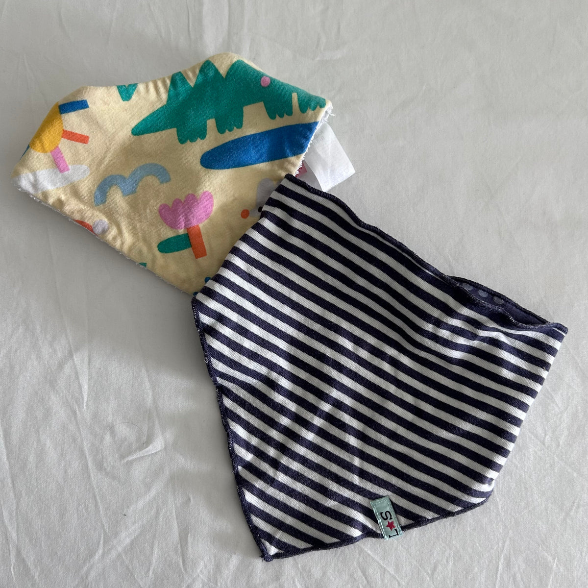 Halcyon Nights + One Other 00 bandana bib set (2 bibs) in cream, teal, yellow, pink, blue, navy, white with crocodile and abstract shapes print on one bib; navy and white stripes on reverse — gently loved condition.