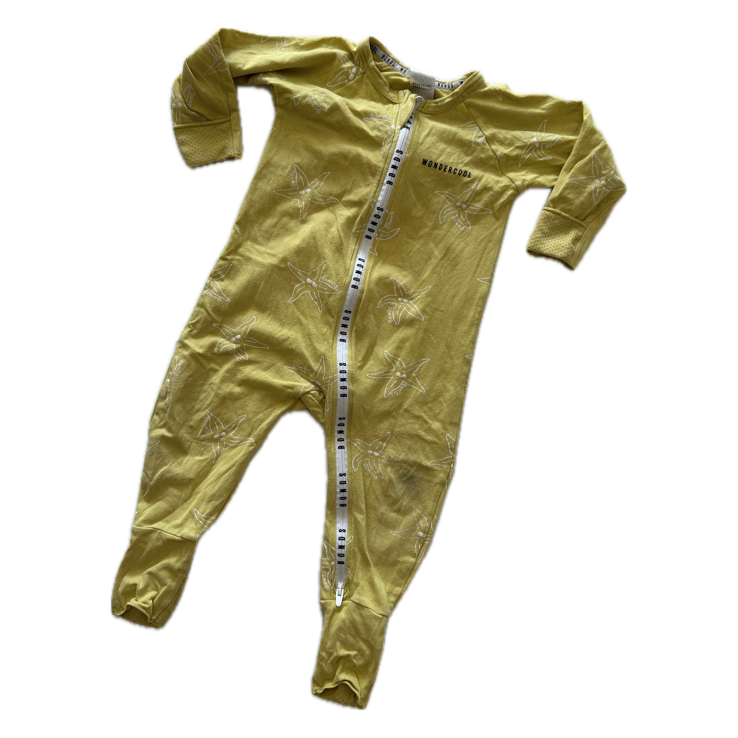 A baby's long-sleeve footed sleeper in a muted mustard yellow color, featuring a subtle tonal palm tree pattern and a white zipper with black Bonds lettering down the front