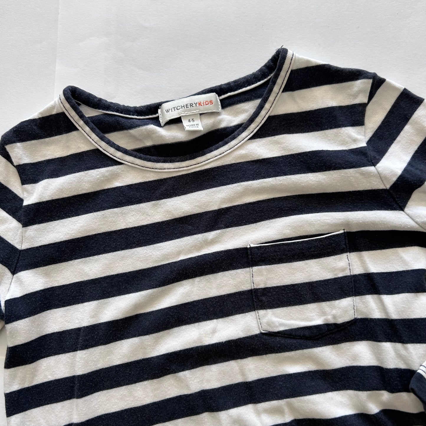 A close-up of a children's long-sleeve striped shirt with a chest pocket, featuring navy blue and off-white horizontal stripes. The brand label 'WITCHERY KIDS' is visible inside the collar.