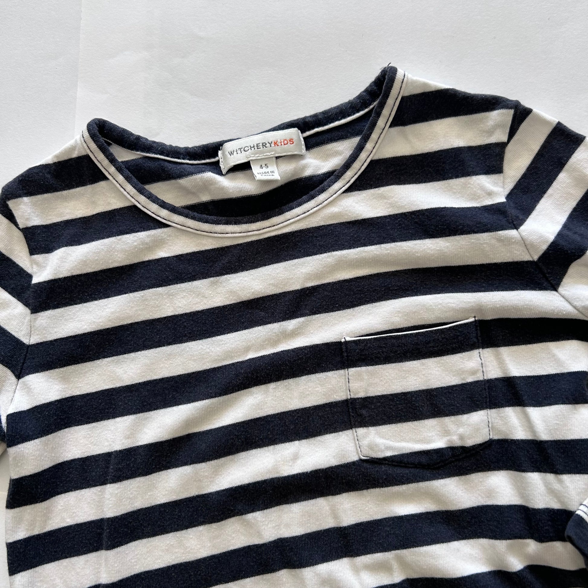 A close-up of a children's long-sleeve striped shirt with a chest pocket, featuring navy blue and off-white horizontal stripes. The brand label 'WITCHERY KIDS' is visible inside the collar.