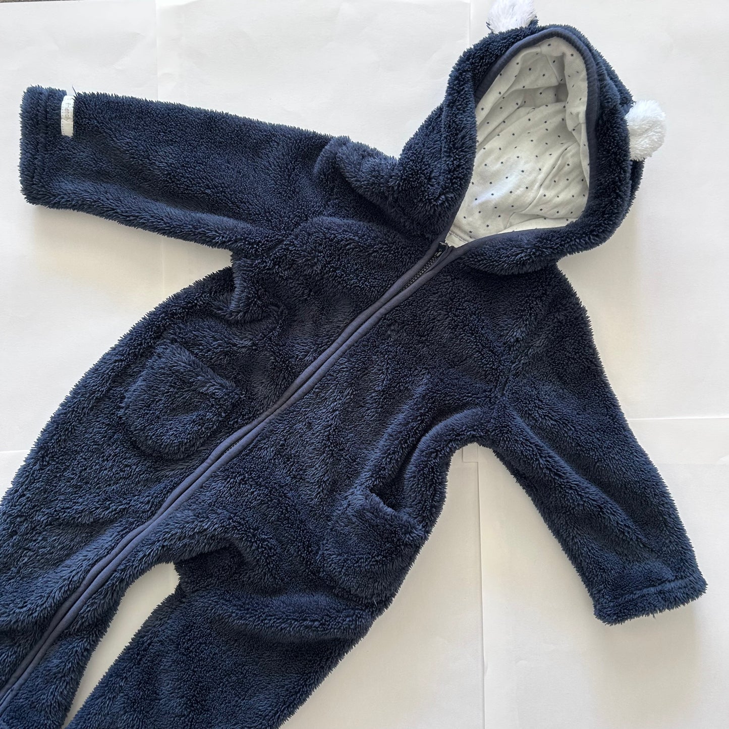 A navy blue plush fleece baby footed one-piece jumpsuit with a front zipper, hood with white pom-poms, and a white patterned lining, laid flat on a white background.