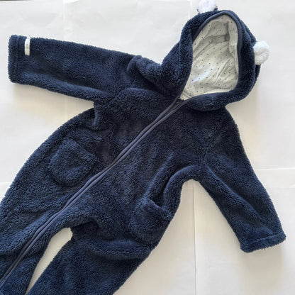 A navy blue plush fleece baby footed one-piece jumpsuit with a front zipper, hood with white pom-poms, and a white patterned lining, laid flat on a white background.