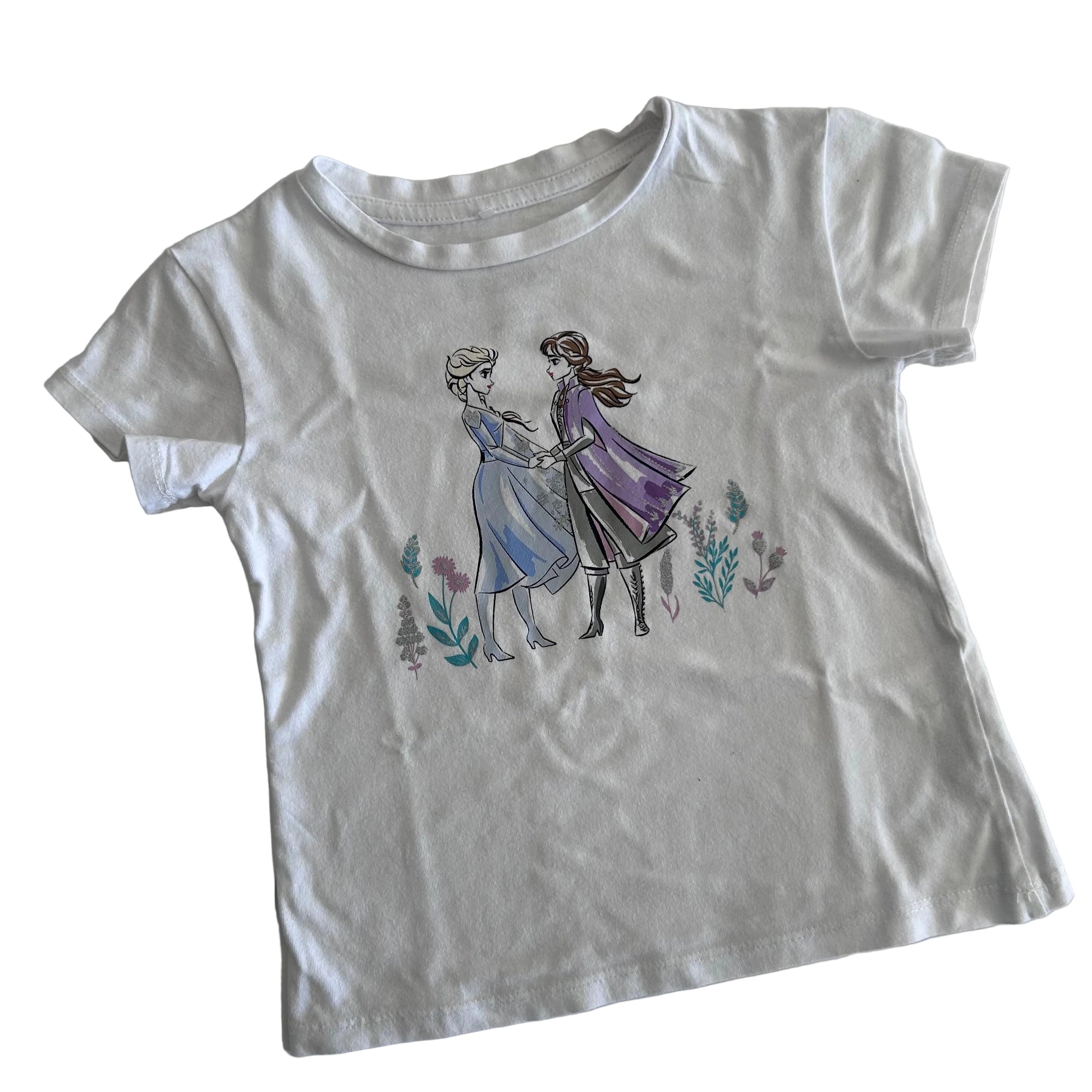 A white short-sleeve kids' t-shirt featuring a printed graphic of Elsa and Anna from Frozen holding hands, surrounded by small watercolor flowers, laid flat on a light background.