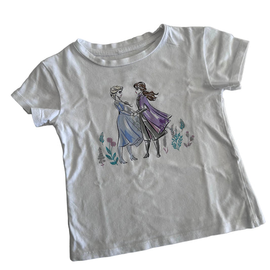 A white short-sleeve kids' t-shirt featuring a printed graphic of Elsa and Anna from Frozen holding hands, surrounded by small watercolor flowers, laid flat on a light background.