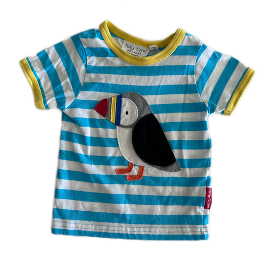 A baby's short-sleeve t-shirt by Toby Tiger, featuring blue and white horizontal stripes with a yellow collar and sleeve trim. The front is decorated with a large, colorful applique puffin.