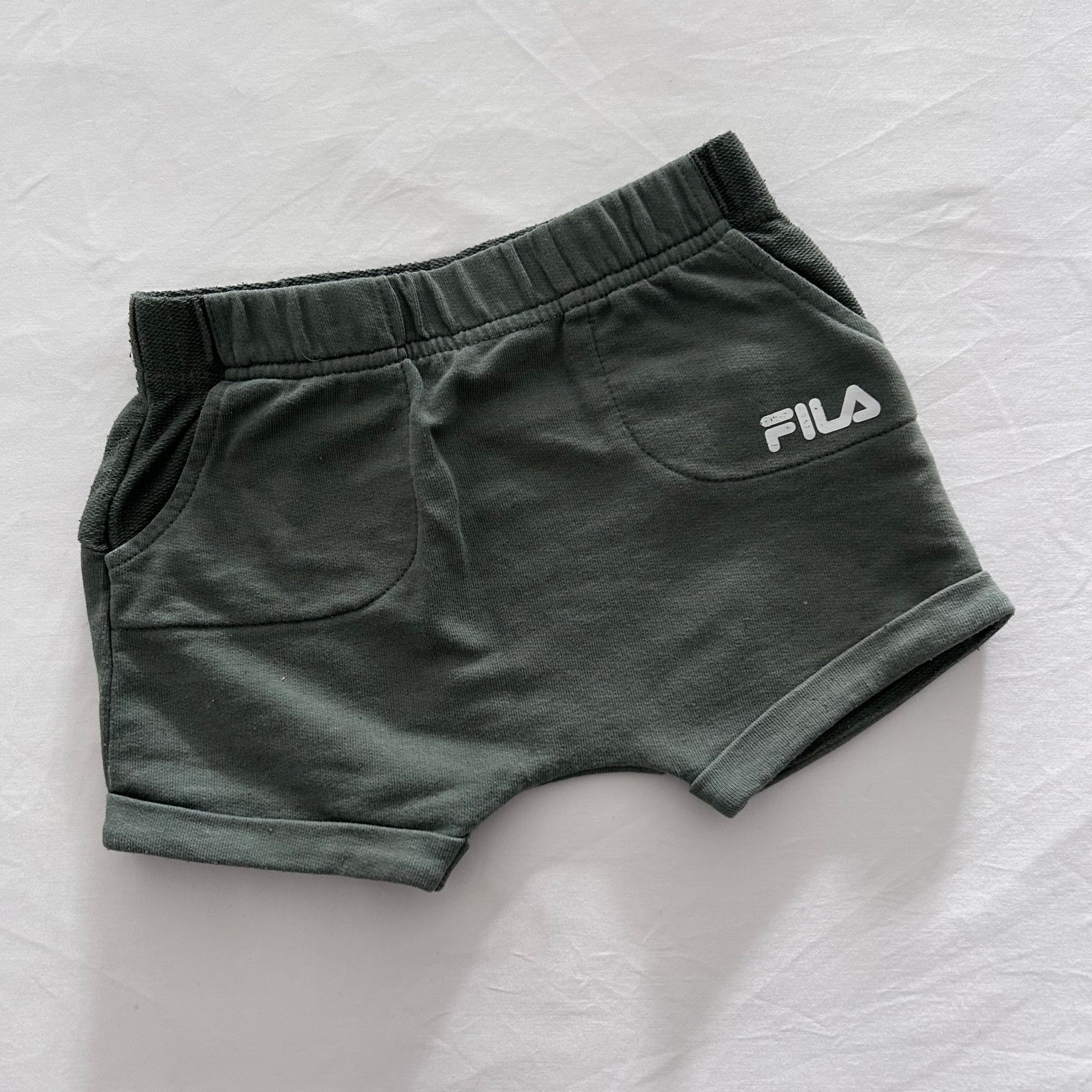 A pair of dark olive green FILA shorts with a white logo on the side, laid flat on a white fabric background.