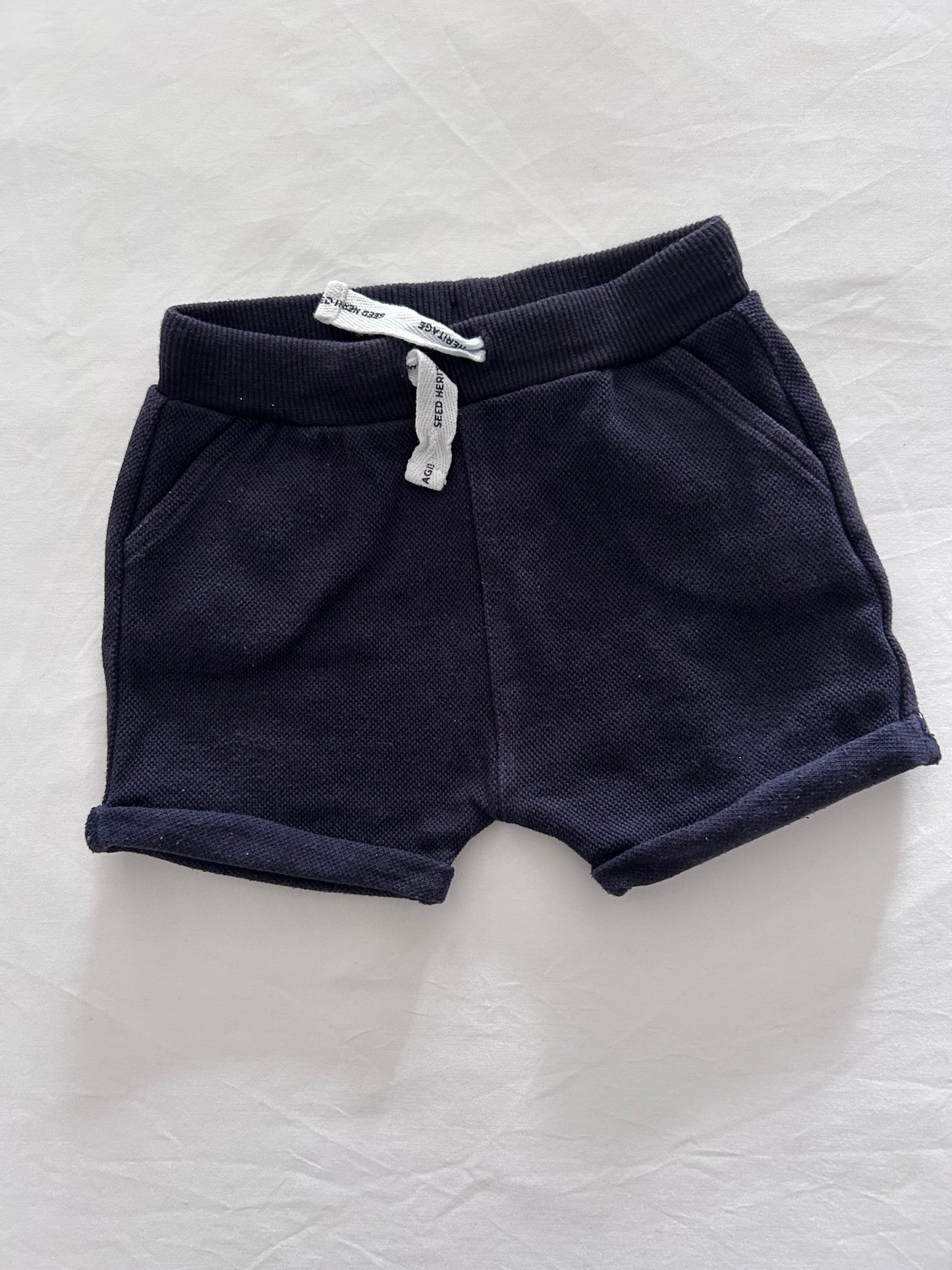 A pair of dark navy blue knit shorts with ribbed cuffs, laid flat on a white textured fabric background. A small white paper tag is attached near the waistband.
