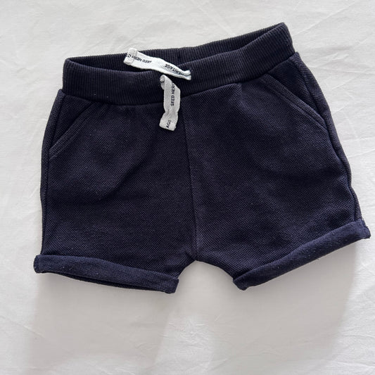 A pair of dark navy blue knit shorts with ribbed cuffs, laid flat on a white textured fabric background. A small white paper tag is attached near the waistband.