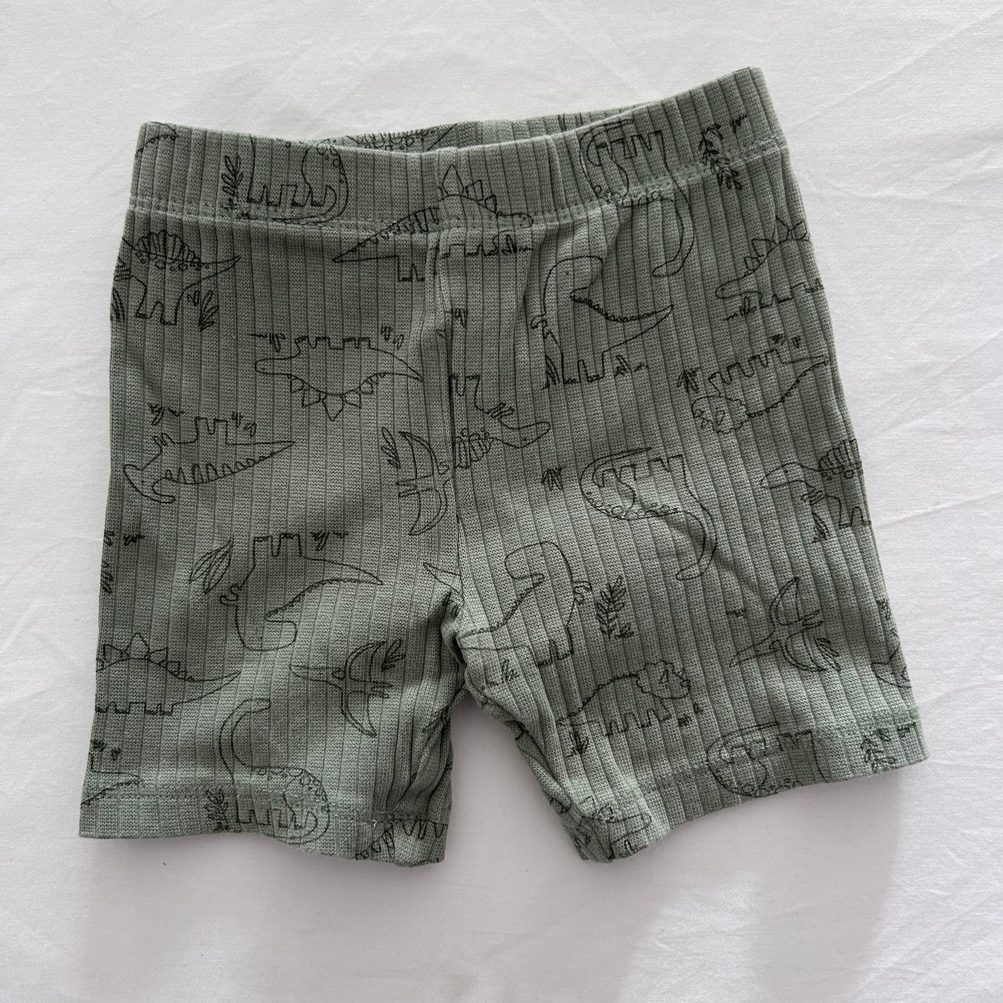 A pair of baby shorts in a muted sage green color, featuring a dark green line art print of various dinosaurs, laid flat on a white fabric background.