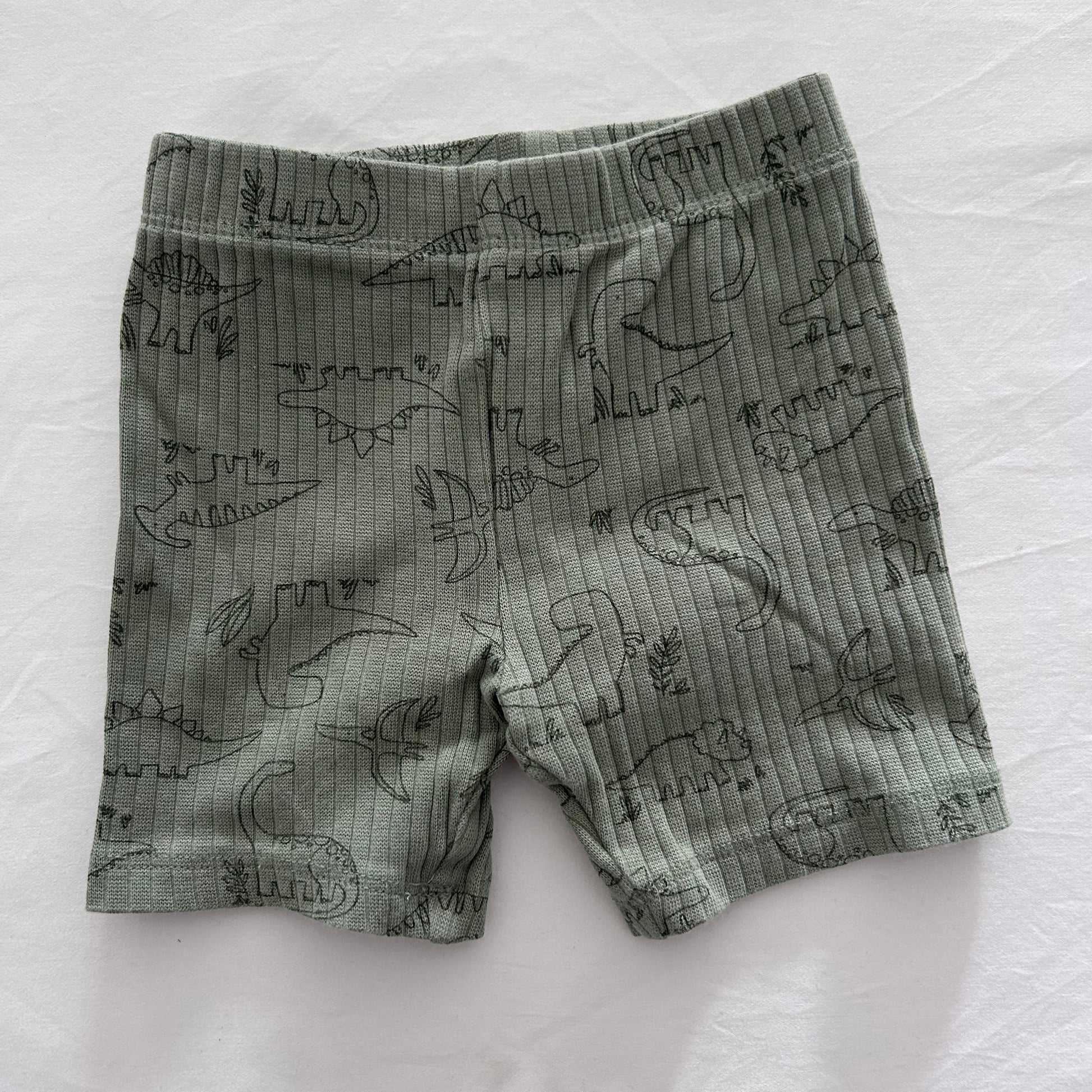 A pair of baby shorts in a muted sage green color, featuring a dark green line art print of various dinosaurs, laid flat on a white fabric background.