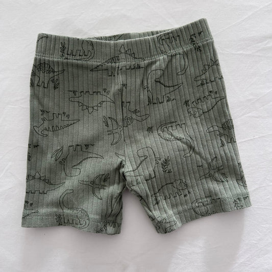 A pair of baby shorts in a muted sage green color, featuring a dark green line art print of various dinosaurs, laid flat on a white fabric background.