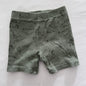 A pair of baby shorts in a muted sage green color, featuring a dark green line art print of various dinosaurs, laid flat on a white fabric background.