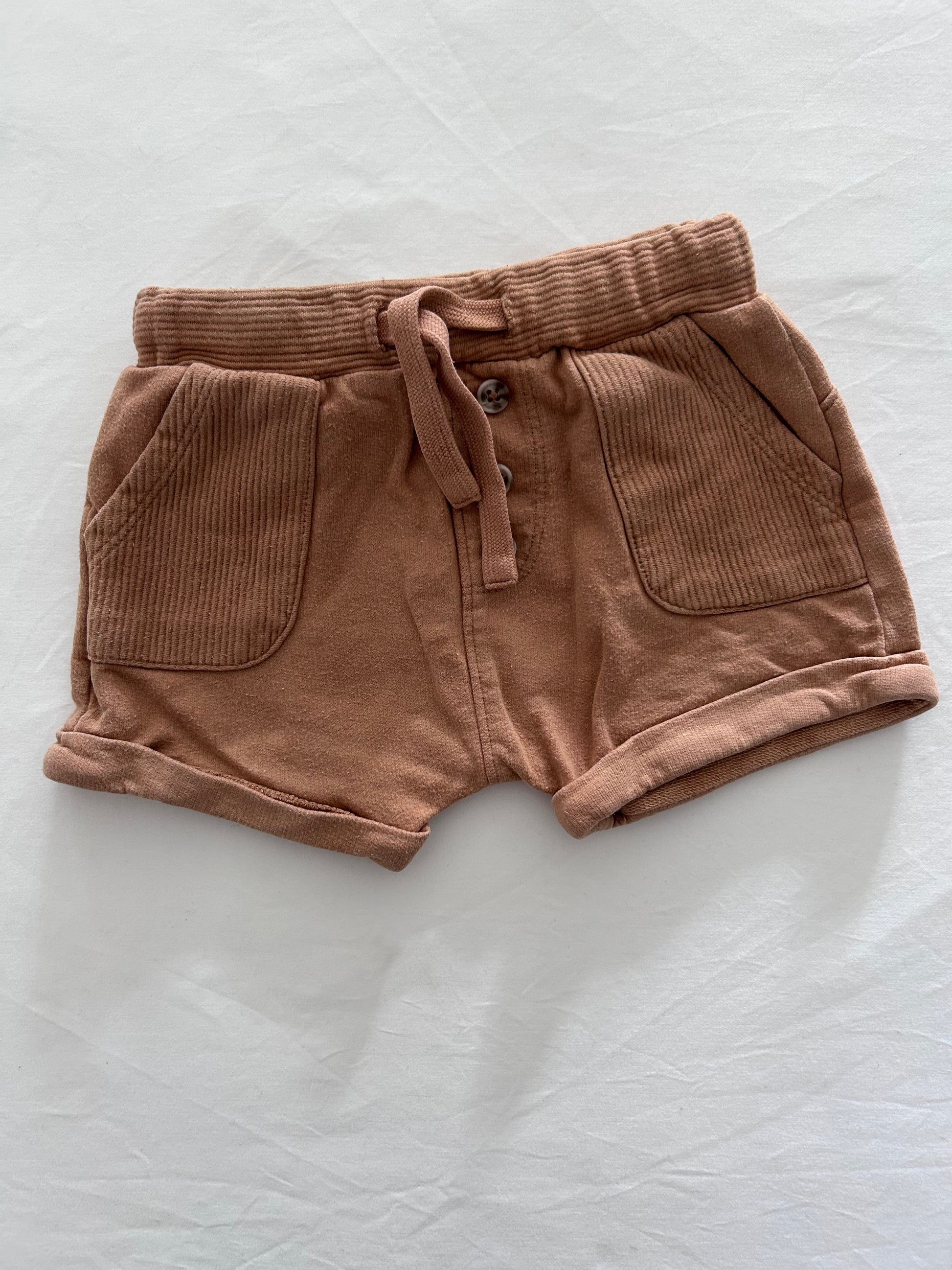 A pair of tan brown shorts with a drawstring waist and front pockets, laid flat on a white fabric background.