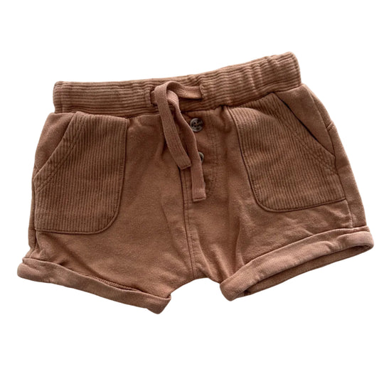 A pair of tan brown shorts with a drawstring waist and front pockets, laid flat on a white fabric background.