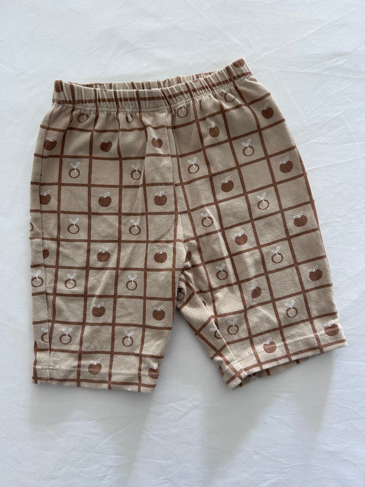 Light brown / tan shorts with dark brown grid lines and pictures of apples in opposite grids, photographed on a white background.