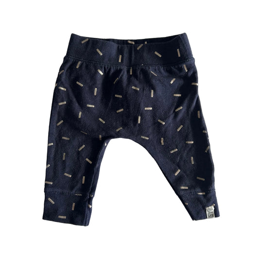 A pair of baby pants in dark navy blue with a scattered beige dash pattern, laid flat on a white fabric background.