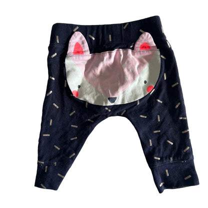 A pair of baby pants in dark navy blue with a white and pink fox face applique on the front and small silver dash patterns scattered across the fabric, laid flat on a white sheet.