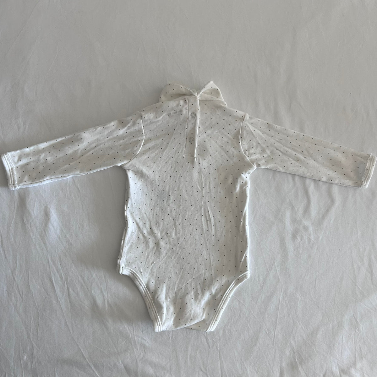 A long-sleeve baby bodysuit in off-white with a tiny dark polka dot pattern, laid flat on a wrinkled white sheet..