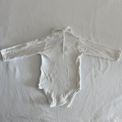 A long-sleeve baby bodysuit in off-white with a tiny dark polka dot pattern, laid flat on a wrinkled white sheet..