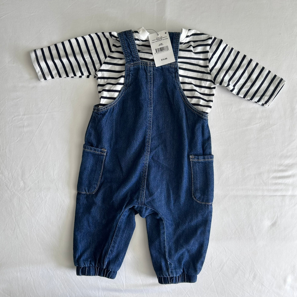 A baby's two-piece outfit featuring blue denim overall pants and a black and white striped long-sleeve shirt, laid flat on a white fabric background with a retail tag attached.