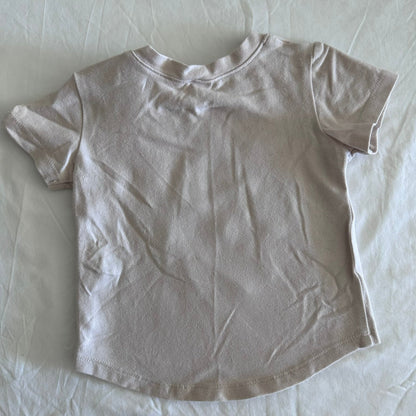 A light beige short-sleeve t-shirt laid flat on a wrinkled white sheet.