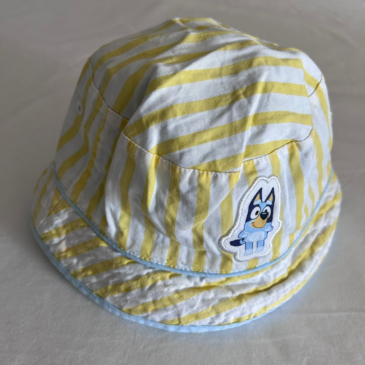 A yellow and white striped baby sun hat with a light blue trim and a Bluey character patch on the side, displayed on a light gray fabric background.