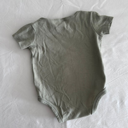A short-sleeve baby bodysuit in a muted sage green color, laid flat on a wrinkled white sheet.