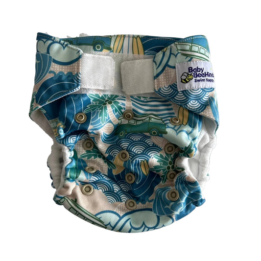 A reusable cloth swim diaper with a blue, green, and beige wave pattern, featuring two white hook-and-loop fastening closures and a white brand label that reads 'Baby Beating Swim Diaper'.
