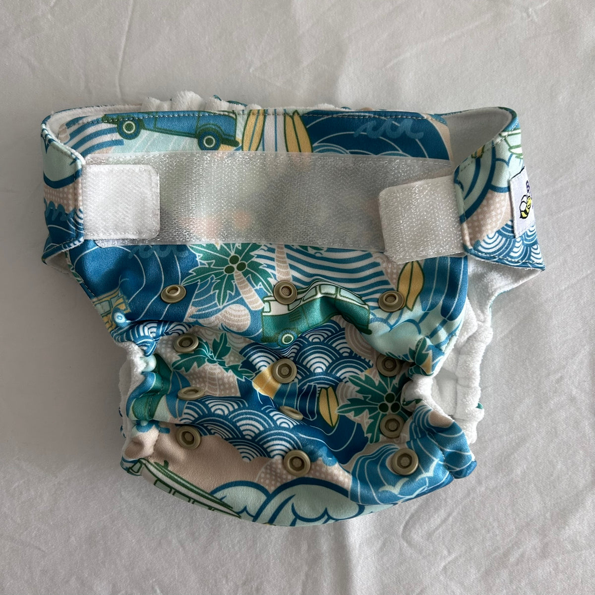 Baby BeeHinds Age 00 swim nappy in blue, teal, white, beige, yellow, green with surf/beach print with waves, palm trees, surfboards and vintage cars — gently loved condition.