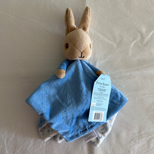 Peter Rabbit security blanket toy with a beige rabbit head, blue fabric body, and attached light blue product tag, photographed on a white fabric surface.