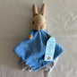 Peter Rabbit security blanket toy with a beige rabbit head, blue fabric body, and attached light blue product tag, photographed on a white fabric surface.