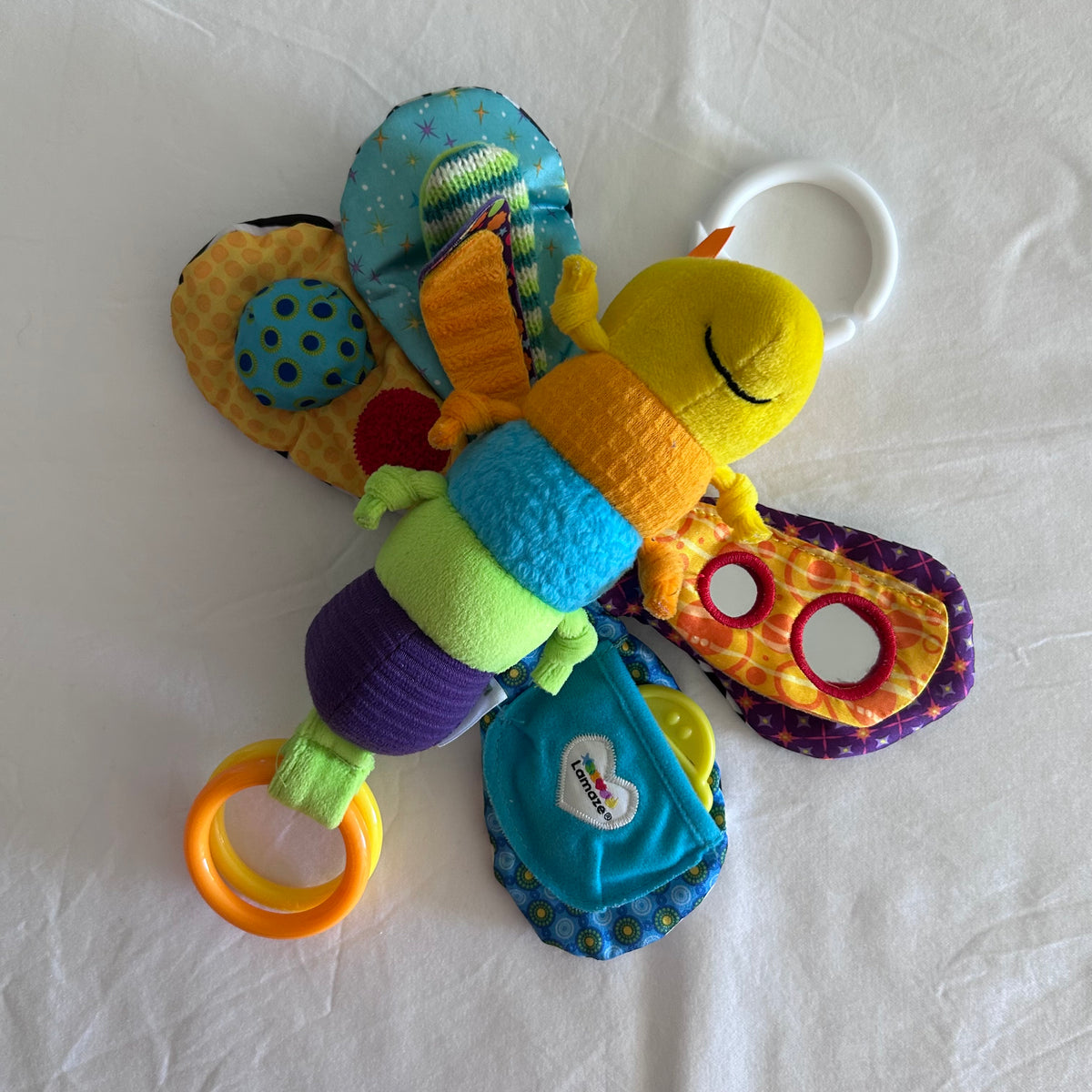 A colorful baby butterfly activity toy with a yellow plush head, patterned wings, and multiple sensory rings and fabric tabs, photographed on a white fabric background.